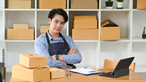 Confident man small business owner wearing apron sitting among cardboard boxes and looking at camera. Online selling, E-commerce concept