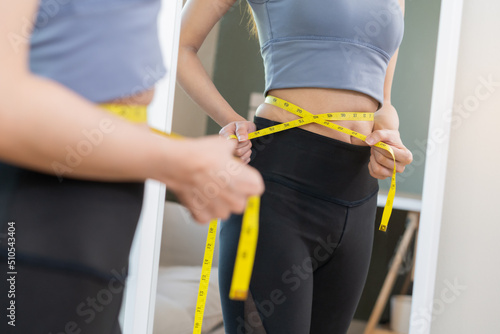 Workout, Slim asian young woman standing hand in measuring tape around waist her in fit sportswear, looking reflect in mirror at home. Sport strong person in gym fitness for wellbeing healthcare.