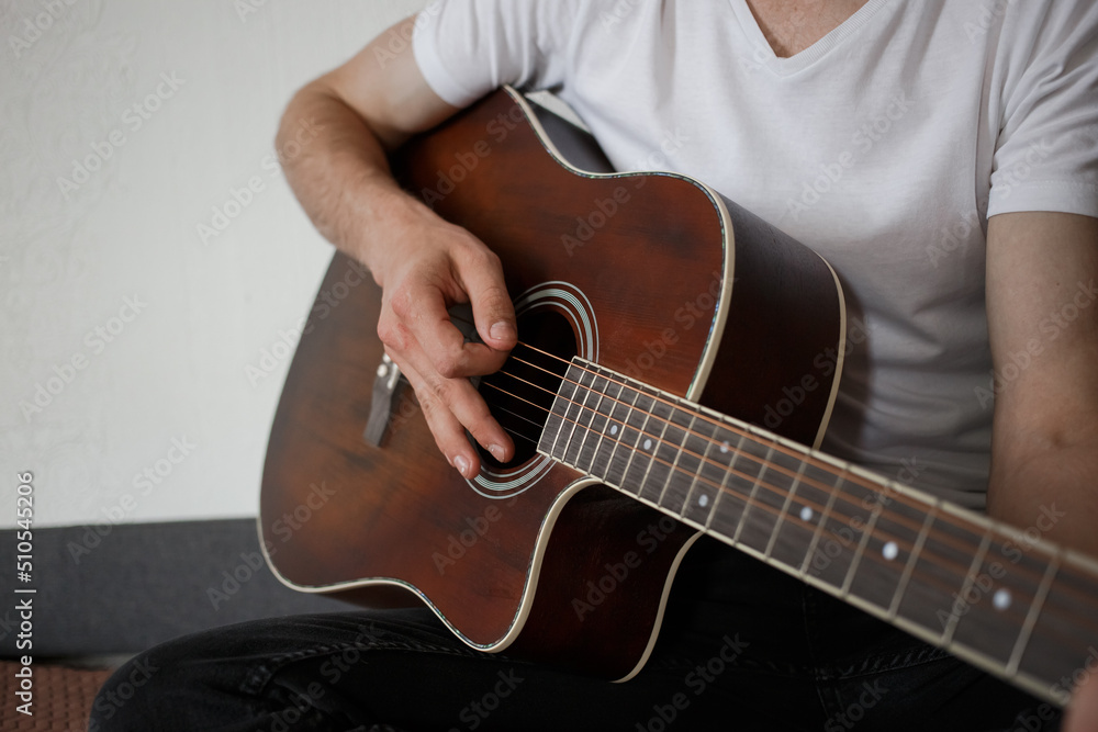 A man in a white T-shirt, playing guitar at home.