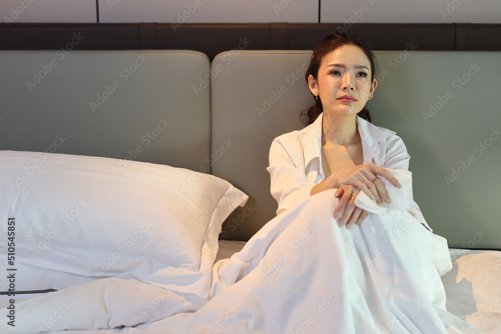 Young depressed asian woman sitting and looking at the ceiling while crying on bed in hotel bedroom with low light environment. Sad girl, unhappy and loneliness concept. Young depressed asian woman sitting and looking at the ceiling while crying on bed in hotel bedroom with low light environment. Sad girl, unhappy and loneliness concept.