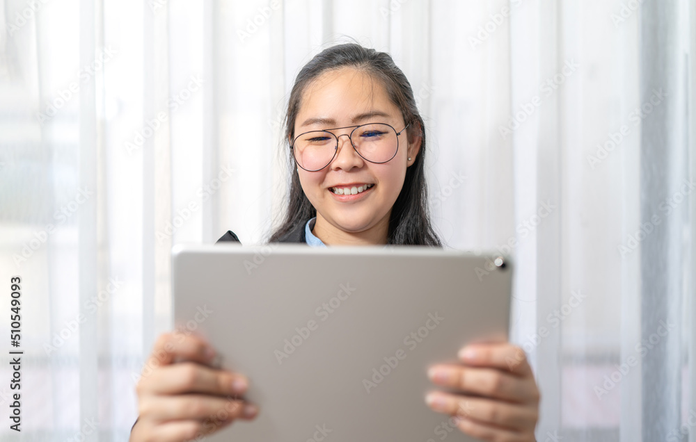 Smiling Asian Woman holds and watches gray tablet on her hand in front of curtain in private room in Work from Home period. Smiling Asian Woman holds and watches gray tablet on her hand in front of curtain in private room in Work from Home period.