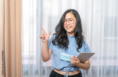 Happy Asian woman fingering up and pointing with tablet computer on her hand at copy space while standing in front of the transparent curtain see through the window light.
