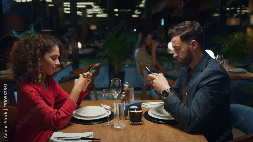 Multiethnic partners searching phone device on fancy restaurant dinner date. 