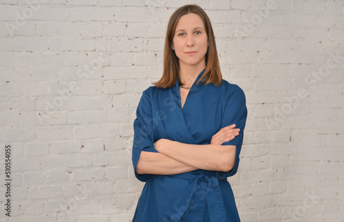 Portrait of middle aged female doctor wearing blue uniform, looking at camera and holding hands crossed on chest on a white brick wall background.

Medical service, medicine or treatment concept. photo