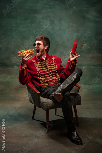 Young man in suit as royal hussar tasting big hot-dog isolated on dark green background. Retro style, comparison of eras concept.