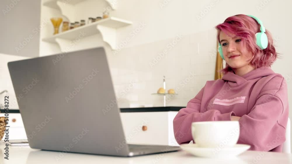 Teen girl student with pink hair wearing headphones looking at laptop computer learning online, watching webinar, remote studying at home. Teen girl student with pink hair wearing headphones looking at laptop computer learning online, watching webinar, remote studying at home.