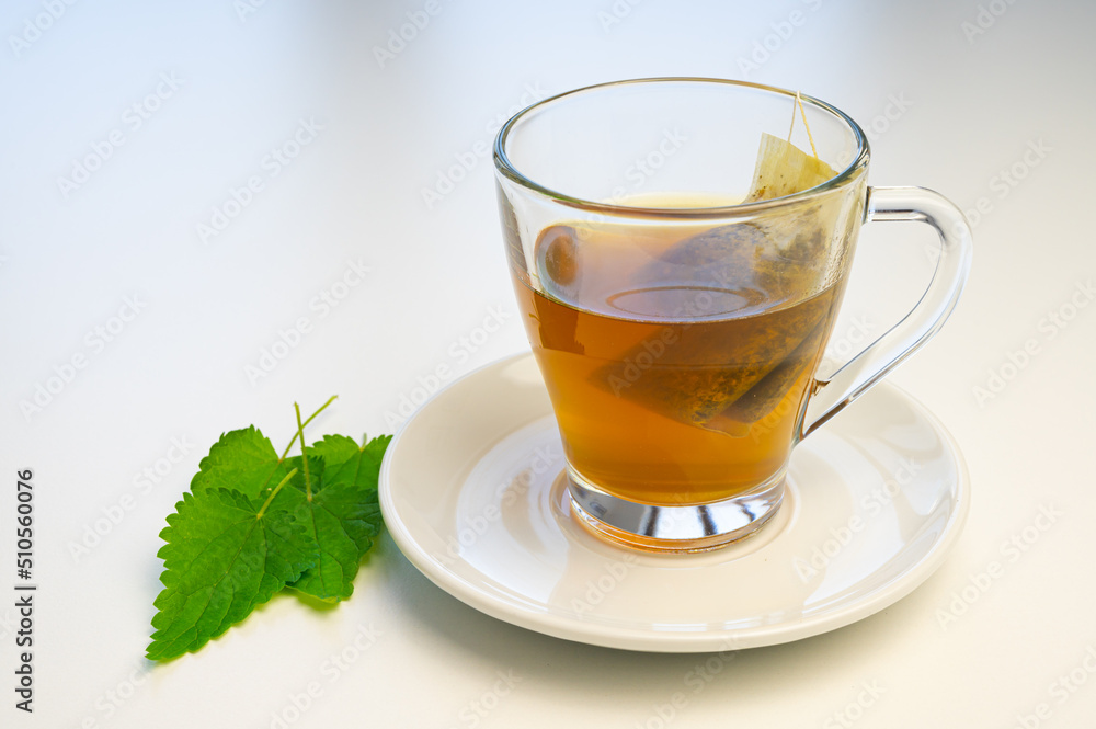 Nettle herbal tea in a transparent cup, with a sachet immersed in water, and a white saucer with nettle leaves next to it. Healing infusion of nettle.
Nettle herbal tea in a transparent cup, with a sachet immersed in water, and a white saucer with nettle leaves next to it. Healing infusion of nettle.