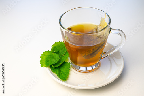 Nettle herbal tea in a transparent cup, with a sachet immersed in water, and a white saucer with nettle leaves next to it. Healing infusion of nettle. 