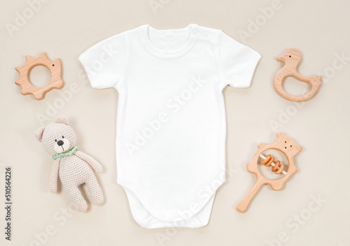White baby bodysuit for mockup on a pastel beige background. Baby wooden teethers and crocketed toy of teddy bear next to white romper for kids clothes mockup.