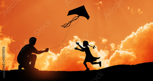 Happy family on the field. Father and daughter playing with a kite while running on meadow on the background of the sunset. Funny family time. Happy little daughter launch a kite with dad.