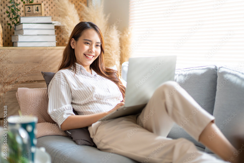 Smiling Asian freelance business women casual wear sitting on sofa using laptop looking at screen typing message,working call video conference in living room.Happy young girl relax at home in summer Smiling Asian freelance business women casual wear sitting on sofa using laptop looking at screen typing message,working call video conference in living room.Happy young girl relax at home in summer