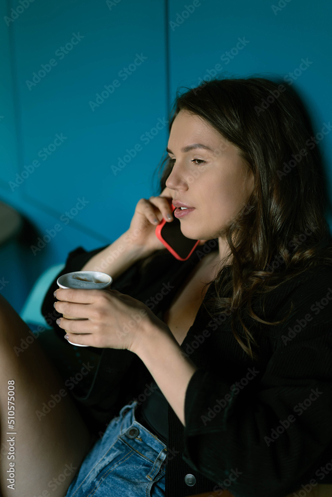 A girl in a cafe is drinking coffee and talking on the phone. Close-up portrait on a blue background. The concept of leisure and wireless technologies . Copy space.