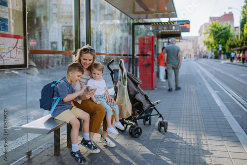 Foto Young mother with little kids waiting on bus stop in city, scrolling on mobile phone