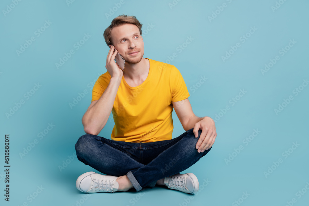 Dreamy positive guy sit floor hold telephone call look empty space wear casual clothes isolated on turquoise background Dreamy positive guy sit floor hold telephone call look empty space wear casual clothes isolated on turquoise background