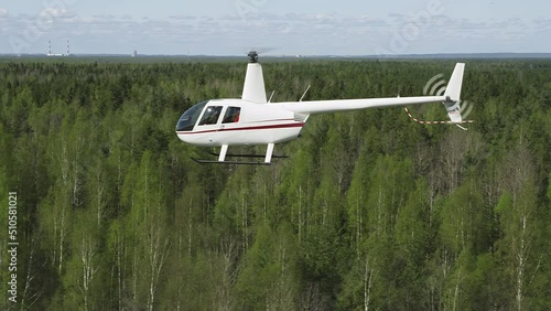 A white Robinson helicopter flies against background of green forest with Christmas trees and a blue sky. Close-up of flying helicopter with an identifying Russian flag on board on sunny summer day photo