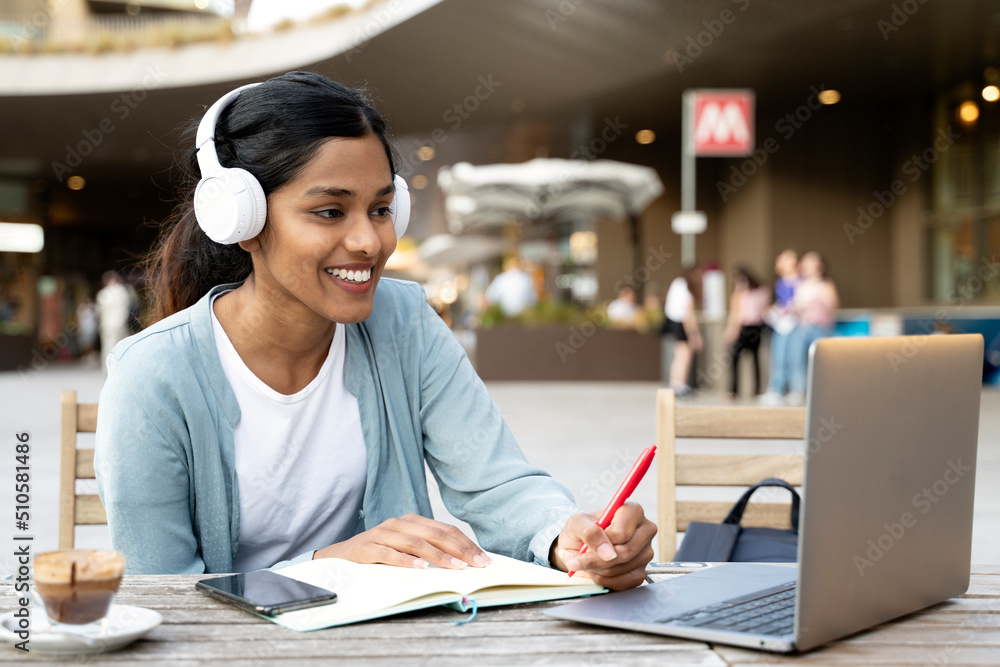 Fototapeta premium Smiling Indian woman using laptop computer studying, taking notes, exam preparation, online education concept. Young asian freelancer working project, watching training courses sitting at workplace