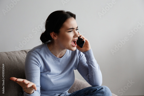 Portrait of irritated young woman arguing on phone. Outraged female talking angrily, shouting at cellphone. Customer support frustration concept. Copy space for text, white wall background, close up.