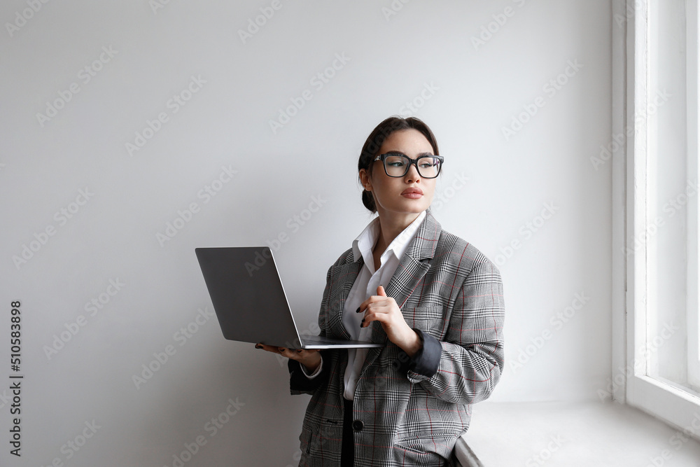 Portrait of a young businesswoman wearing smart casual attire standing with a laptop. Studio shot of a brunette female in eyeglasses posing near white wall. Close up, copy space, background. Portrait of a young businesswoman wearing smart casual attire standing with a laptop. Studio shot of a brunette female in eyeglasses posing near white wall. Close up, copy space, background.