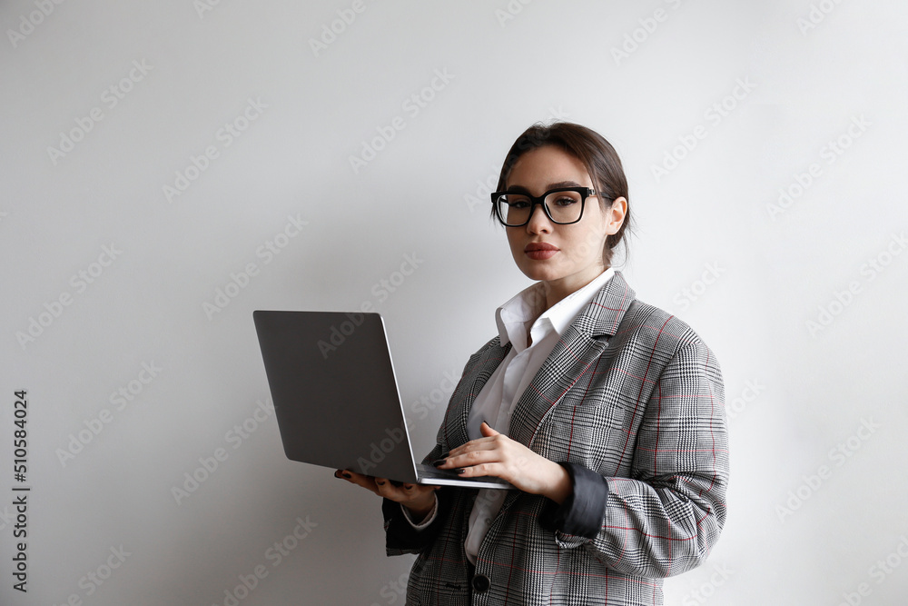Portrait of a young businesswoman wearing smart casual attire standing with a laptop. Studio shot of a brunette female in eyeglasses posing near white wall. Close up, copy space, background. Portrait of a young businesswoman wearing smart casual attire standing with a laptop. Studio shot of a brunette female in eyeglasses posing near white wall. Close up, copy space, background.
