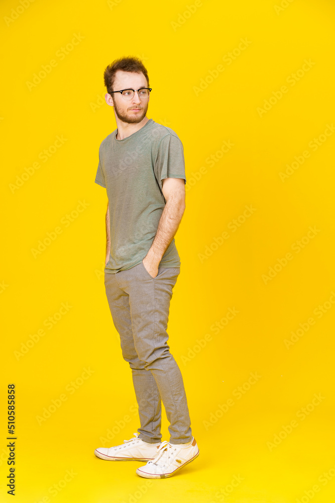 Young man wears eye glasses in casual wear posing on yellow background with hands in pockets looking sideways. Stylish bearded smart casual look hipster man. Thoughtful guy on yellow background Young man wears eye glasses in casual wear posing on yellow background with hands in pockets looking sideways. Stylish bearded smart casual look hipster man. Thoughtful guy on yellow background