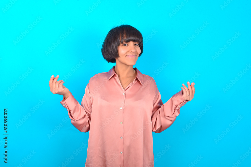 young brunette woman wearing pink silk shirt over blue background, making money gesture. young brunette woman wearing pink silk shirt over blue background, making money gesture.