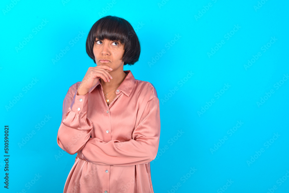 Shot of contemplative thoughtful young brunette woman wearing pink silk shirt over blue background keeps hand under chin, looks thoughtfully upwards. Shot of contemplative thoughtful young brunette woman wearing pink silk shirt over blue background keeps hand under chin, looks thoughtfully upwards.