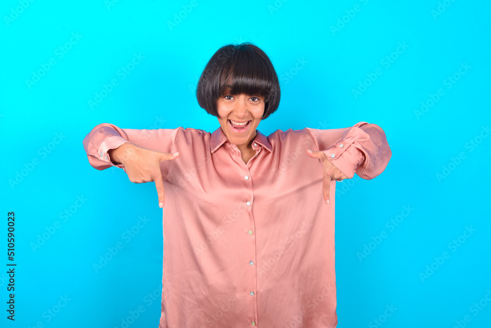 young brunette woman wearing pink silk shirt over blue background with positive expression, points down with both index fingers, has broad interested smile. Look there, please. young brunette woman wearing pink silk shirt over blue background with positive expression, points down with both index fingers, has broad interested smile. Look there, please.