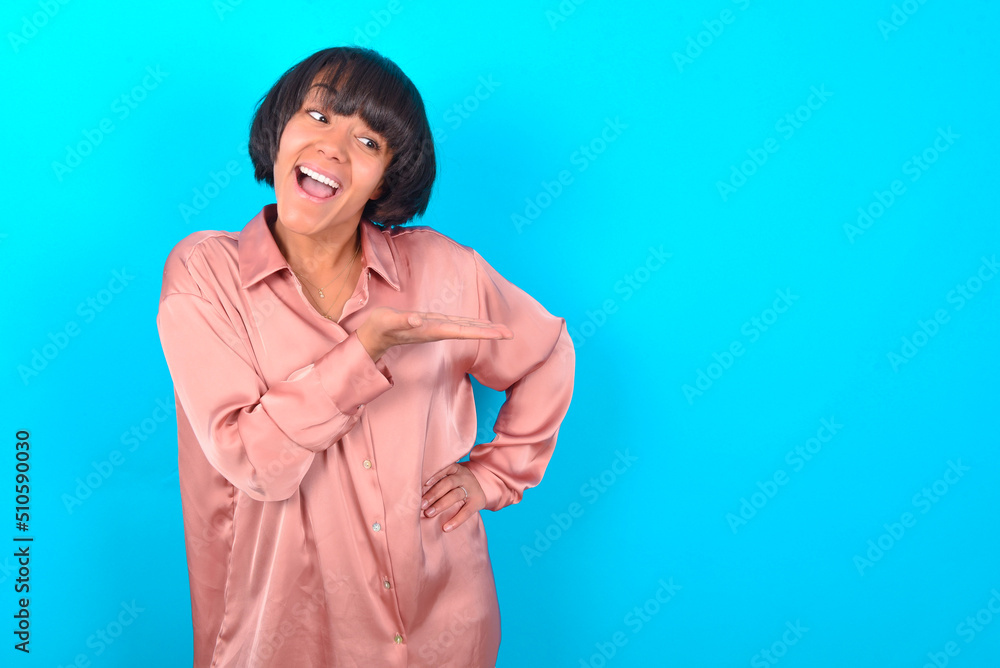 young brunette woman wearing pink silk shirt over blue background says: wow how exciting it is, has amazed expression, shows something on blank space with palm. Advertisement concept. young brunette woman wearing pink silk shirt over blue background says: wow how exciting it is, has amazed expression, shows something on blank space with palm. Advertisement concept.