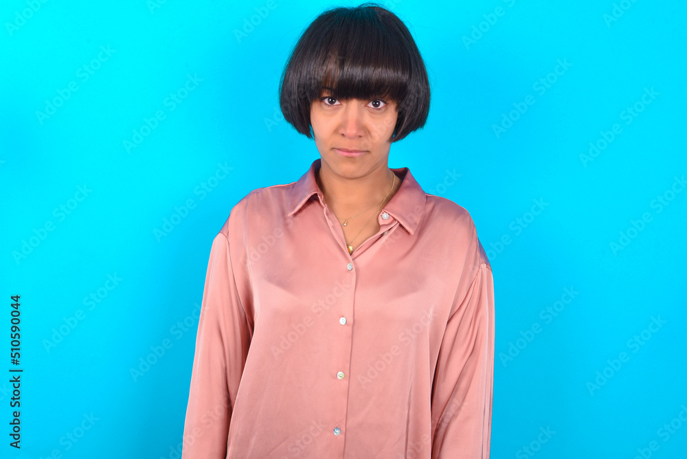 young brunette woman wearing pink silk shirt over blue background frowning his eyebrows being displeased with something. young brunette woman wearing pink silk shirt over blue background frowning his eyebrows being displeased with something.