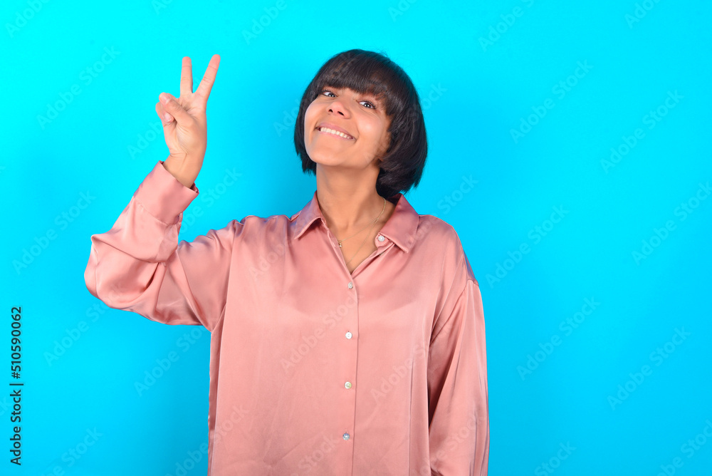 young brunette woman wearing pink silk shirt over blue background showing and pointing up with fingers number two while smiling confident and happy. young brunette woman wearing pink silk shirt over blue background showing and pointing up with fingers number two while smiling confident and happy.