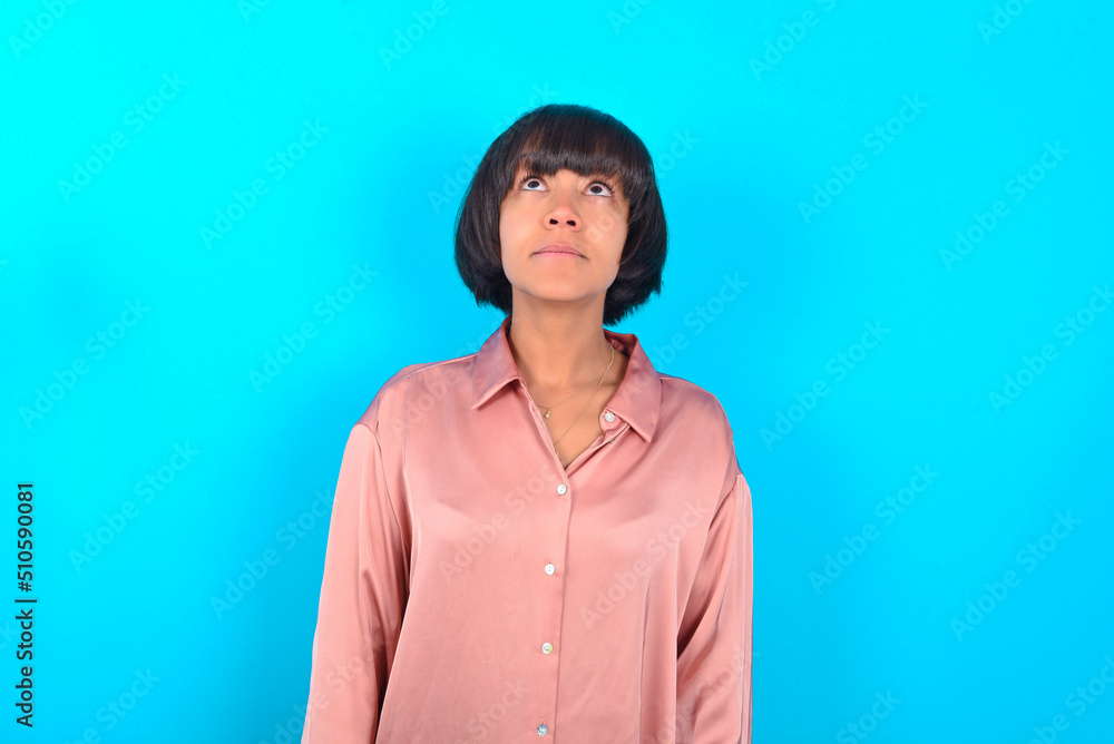 Fototapeta premium young brunette woman wearing pink silk shirt over blue background looking up as he sees something strange.