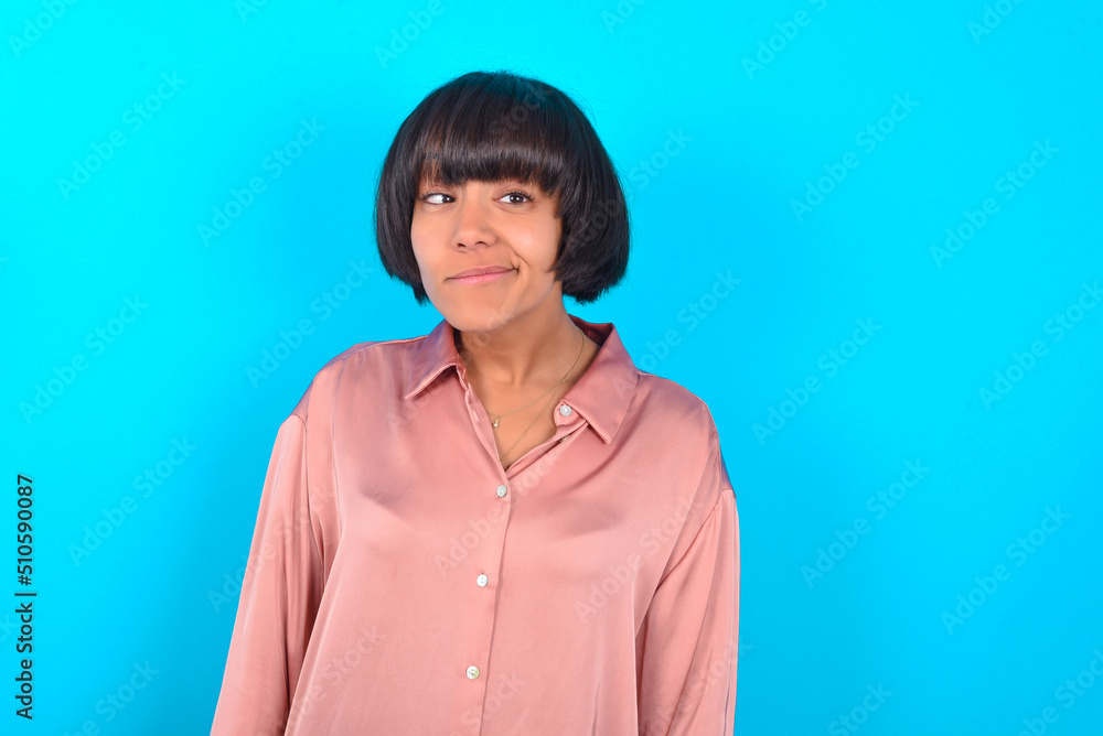 young brunette woman wearing pink silk shirt over blue background has worried face looking up lips together, being upset thinking about something important, keeps hands down. young brunette woman wearing pink silk shirt over blue background has worried face looking up lips together, being upset thinking about something important, keeps hands down.