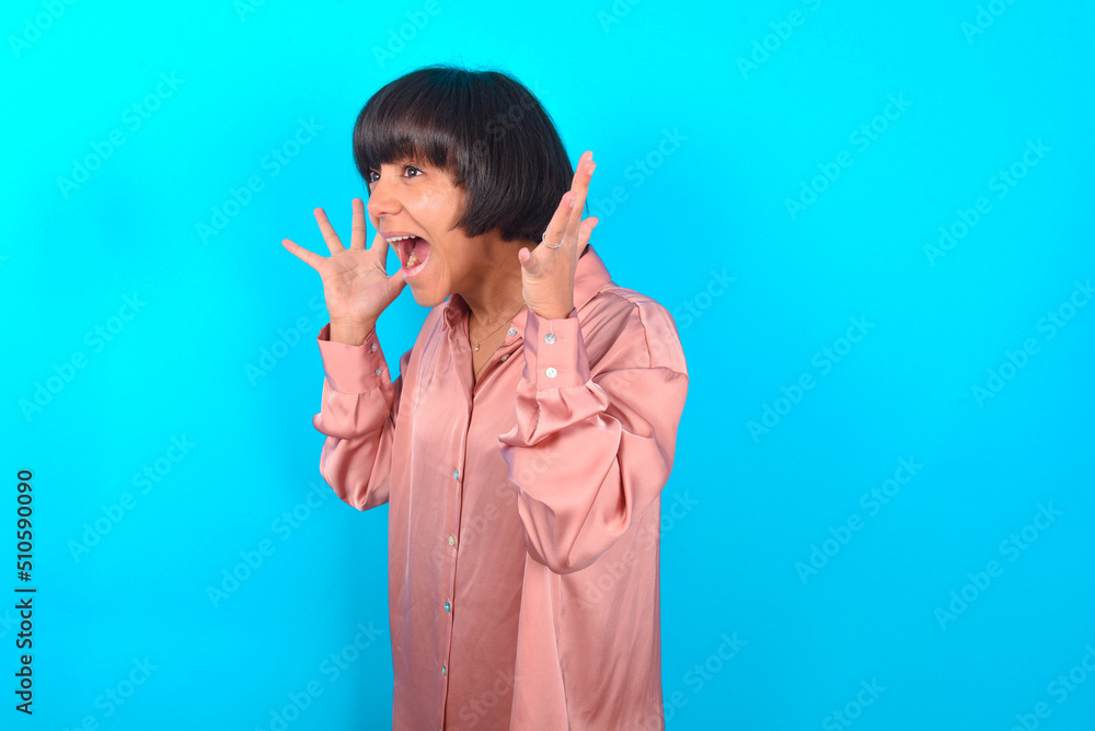 young brunette woman wearing pink silk shirt over blue background with shocked facial expression, holding hands near face, screaming and looking sideways at something amazing. young brunette woman wearing pink silk shirt over blue background with shocked facial expression, holding hands near face, screaming and looking sideways at something amazing.
