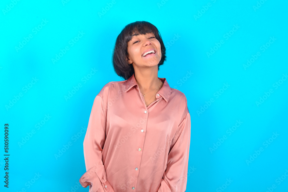 young brunette woman wearing pink silk shirt over blue background with broad smile, shows white teeth, feeling confident rejoices having day off. young brunette woman wearing pink silk shirt over blue background with broad smile, shows white teeth, feeling confident rejoices having day off.