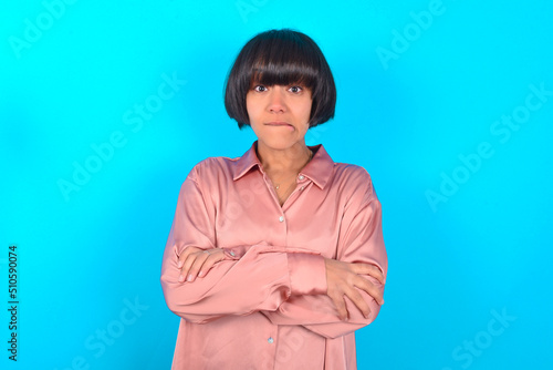 young brunette woman wearing pink silk shirt over blue background bitting his mouth and looking worried and scared crossing arms, worry and doubt.