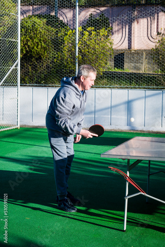 An elderly, gray-haired retired man plays table tennis ping pong on an outdoor sports field on a sunny day. Motion blur on the ball. The concept of an active lifestyle of elderly pensioners