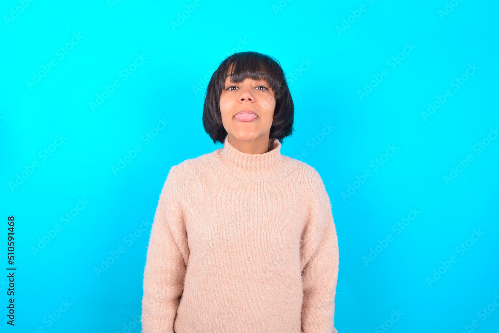 young brunette woman wearing pink knitted sweater over blue background with happy and funny face smiling and showing tongue. young brunette woman wearing pink knitted sweater over blue background with happy and funny face smiling and showing tongue.
