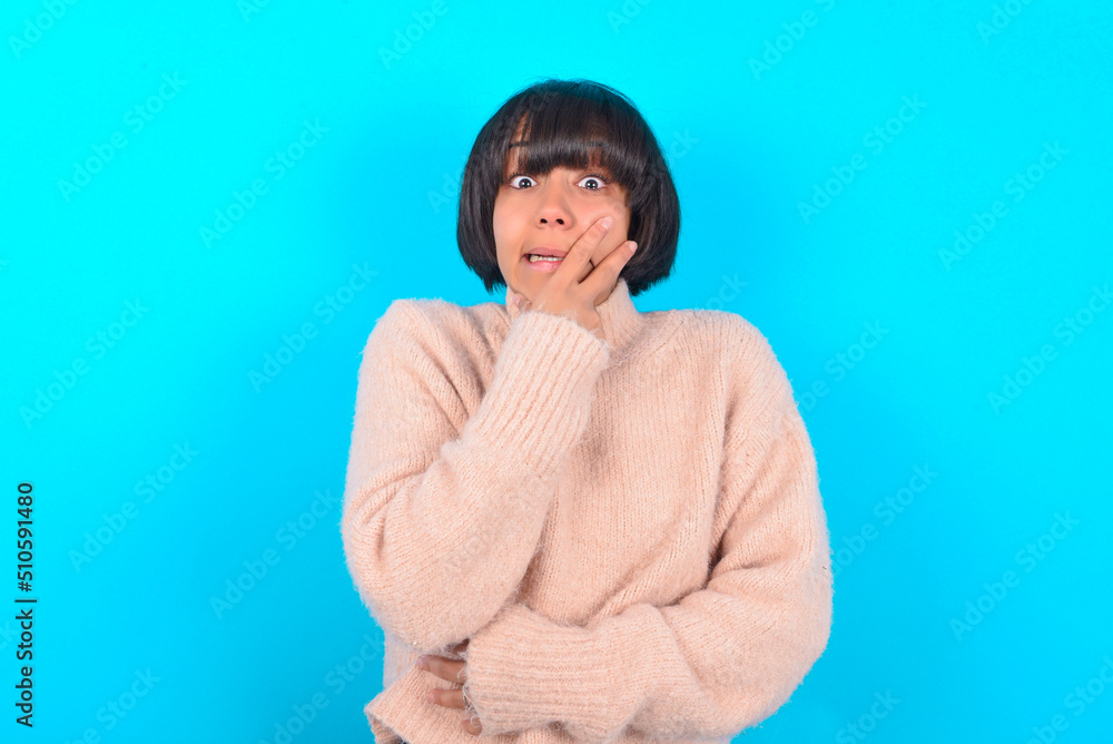 young brunette woman wearing pink knitted sweater over blue background covering mouth with hands scared from something or someone bitting nails young brunette woman wearing pink knitted sweater over blue background covering mouth with hands scared from something or someone bitting nails