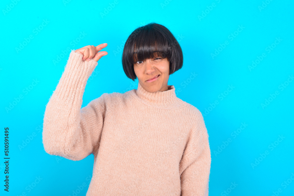 Upset young brunette woman wearing pink knitted sweater shirt over blue background shapes little gesture with hand demonstrates something very tiny small size. Not very much Upset young brunette woman wearing pink knitted sweater shirt over blue background shapes little gesture with hand demonstrates something very tiny small size. Not very much