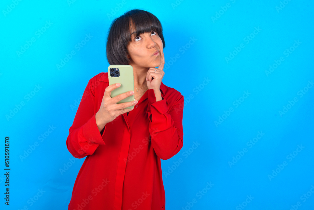 Fototapeta premium Thinking dreaming young brunette woman with short hair wearing red shirt over blue background using mobile phone and holding hand on face. Taking decisions and social media concept.