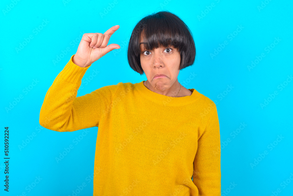 young brunette woman with short hair wearing yellow knitted sweater over blue background purses lip and gestures with hand, shows something very little. young brunette woman with short hair wearing yellow knitted sweater over blue background purses lip and gestures with hand, shows something very little.