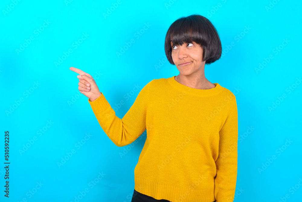 young brunette woman with short hair wearing yellow knitted sweater over blue background points at copy space and advertises something, advices best price. young brunette woman with short hair wearing yellow knitted sweater over blue background points at copy space and advertises something, advices best price.