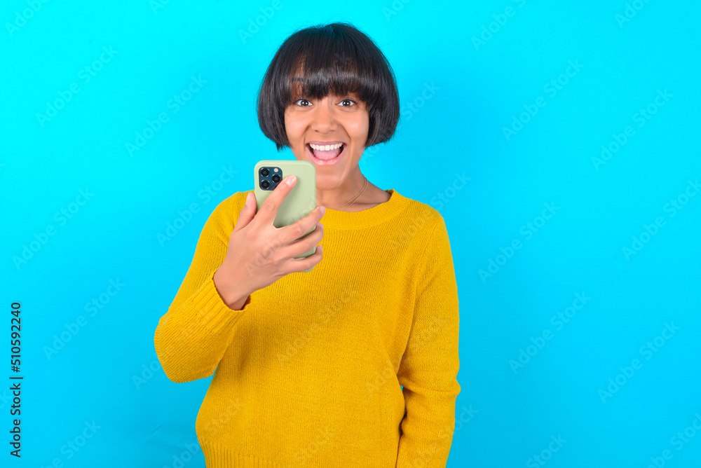 young brunette woman with short hair wearing yellow knitted sweater over blue background holds mobile phone in hands and rejoices positive news, uses modern cellular young brunette woman with short hair wearing yellow knitted sweater over blue background holds mobile phone in hands and rejoices positive news, uses modern cellular