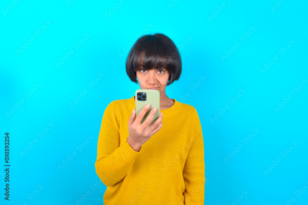 Upset dissatisfied young brunette woman with short hair wearing yellow knitted sweater over blue background uses mobile software application and surfs information in internet, holds modern mobile hand Upset dissatisfied young brunette woman with short hair wearing yellow knitted sweater over blue background uses mobile software application and surfs information in internet, holds modern mobile hand