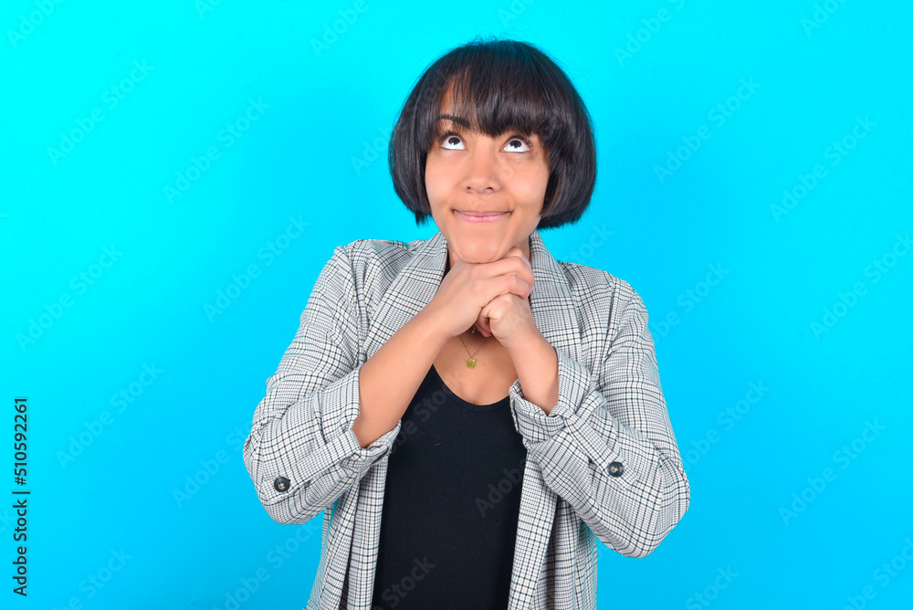 Happy young businesswoman with bob haircut wearing blazer against blue wall anticipates something awesome happen, looks happily aside, keeps hands together near face, has glad expression. Happy young businesswoman with bob haircut wearing blazer against blue wall anticipates something awesome happen, looks happily aside, keeps hands together near face, has glad expression.