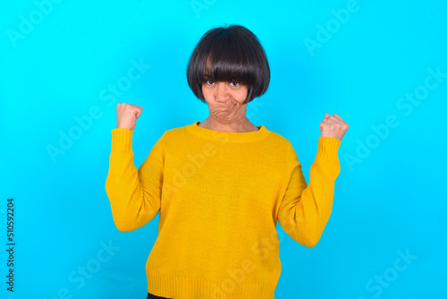 Irritated young brunette woman with short hair wearing yellow knitted sweater over blue backgrou blows cheeks with anger and raises clenched fists expresses rage and aggressive emotions. Furious model