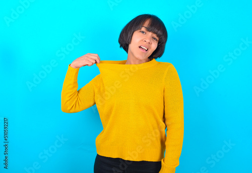 young brunette woman with short hair wearing yellow knitted sweater over blue background stressed, anxious, tired and frustrated, pulling shirt neck, looking frustrated with problem