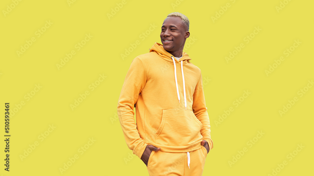 Obraz premium Young black man feeling happy, positive and successful, hands in pocket, achievements or good luck against yellow wall. African ethnicity male person yellow hair yellow hoodie