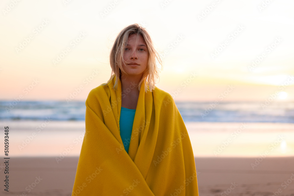 Beautiful young woman wrapped in yellow blanket standing on sandy beach at sunset looking camera. Beautiful caucasian female person wrapped cozy plaid standing alone on ocean beach Beautiful young woman wrapped in yellow blanket standing on sandy beach at sunset looking camera. Beautiful caucasian female person wrapped cozy plaid standing alone on ocean beach