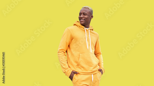 Young black man feeling happy, positive and successful, hands in pocket, achievements or good luck against yellow wall. African ethnicity male person yellow hair yellow hoodie